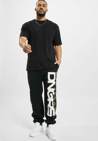 Schwarzes Kurzarm-T-Shirt und schwarze Jogginghosen mit großen weißen "DNGRS"-Schriftzug und gelben Akzenten. Weiße Sneaker runden das Outfit ab.
