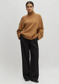 Brauner übergroßer Rollkragensweater mit rippthem, kombiniert mit schwarzen weit geschnittenen Hosen und schwarzen Slippern, vor einem neutralen Hintergrund.