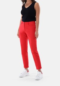 Czerwone, dopasowane spodnie wykonane z gładkiego materiału, z przodem zapinanym na guzik i podwiniętymi mankietami, w zestawieniu z białymi trampkami.
