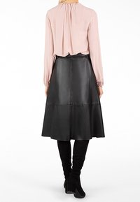 Femme portant un chemisier rose pâle à manches longues, une jupe midi en cuir noire en ligne A et des bottes noires à talons hautes jusqu'aux genoux, vue de dos.