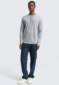 Szara, długosleeve koszula henley z trzema czarnymi guzikami przy kołnierzyku, w połączeniu z niebieskimi dżinsami i białymi trampkami, stojąca w wyprostowanej pozycji.