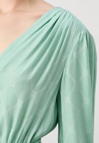 Mintgroene stof met subtiel geometrisch patroon en "HUGO" tekst, met een V-hals en geplooide schouderdetail op een lang mouw kledingstuk.