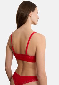 Frau von hinten gesehen, trägt roten BH mit Spitzenbesatz und passende Unterwäsche vor einfachem hellem Hintergrund.