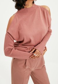 Lys rosa farvet sweater med ribbet høj hals, åbne skulderudskæringer og lange ærmer. Blød tekstur, parret med matchende bukser.