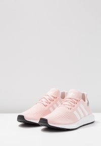 Vaaleanpunaiset urheilukengät verkkomateriaalista valmistetulla päällisellä, valkoisilla raidoilla, teksturoidulla kankaalla ja mustalla ulkopohjalla. Nauhat ja pehmustettu kaulus viimeistelevät mallin.