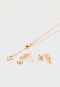 Σετ χρυσών κοσμημάτων που περιλαμβάνει ένα λεπτό κολιέ με αλυσίδα και τρίγωνο μενταγιόν, καθώς και ταιριαστά σκουλαρίκια με περίτεχνα σχέδια κόμπων.