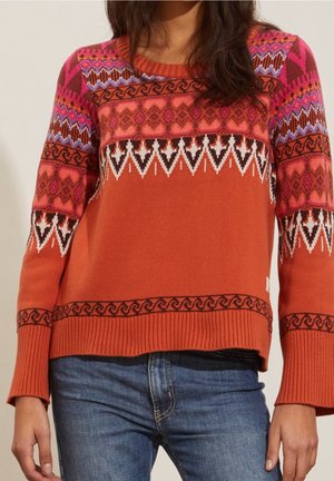 Femme portant un pull orange rouille avec des motifs géométriques multicolores et en zigzag, associé à un jean en denim bleu, debout devant un fond uni.