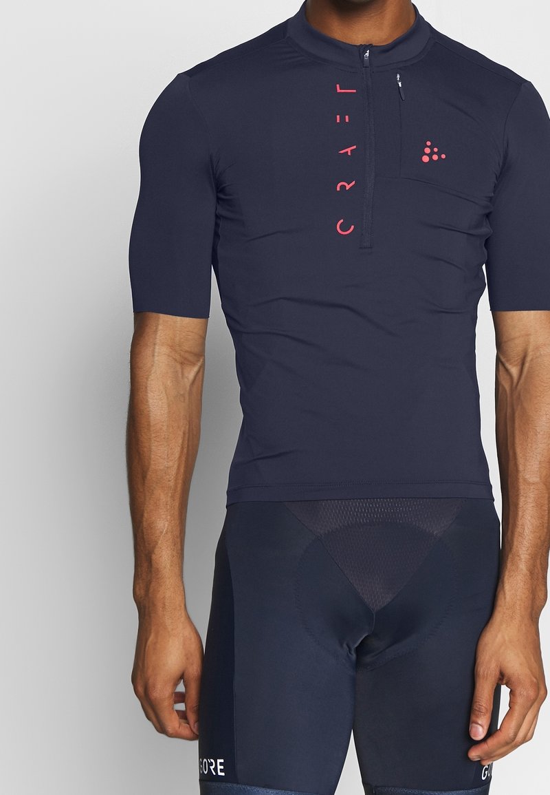 Homme portant un maillot de cyclisme bleu marine à manches courtes avec demi-fermeture éclair et texte vertical rose "CRAFT" à l’avant.