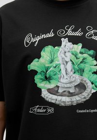 Camiseta negra con un gráfico de una fuente de piedra con un querubín, rodeada de hojas verdes. El texto incluye "Originals Studio" y "Atelier '90."