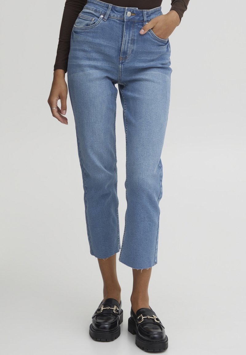 Femme portant un jean court bleu clair avec des ourlets bruts et des mocassins noirs avec des détails de boucle dorée, debout avec une main dans la poche.