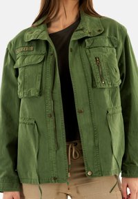 Chaqueta verde de estilo militar con múltiples bolsillos, cierres de cremallera y botón, y cordones ajustables en el dobladillo. Tela de algodón con un acabado texturizado.