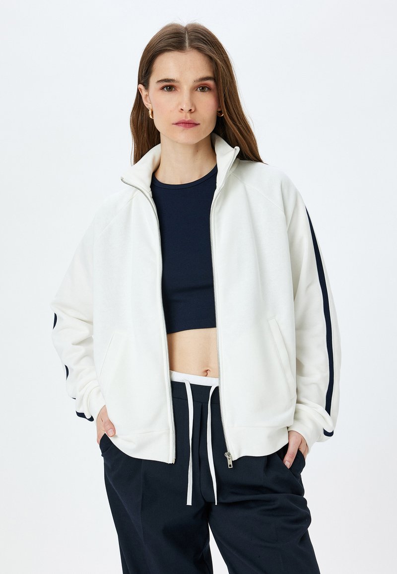 Femme portant une veste blanche zippée avec des rayures bleues sur les manches, un crop top bleu marine et un pantalon bleu marine à cordon de serrage, se tenant avec les mains dans les poches.