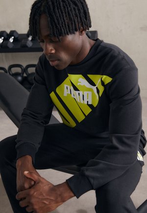 Felpa nera con una grafica gialla in evidenza e testo "PUMA". Design elegante con polsini a coste, abbinata a pantaloni neri coordinati.