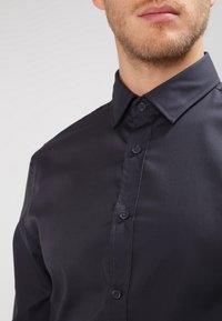 Camicia blu navy con bottoni, realizzata in tessuto liscio; presenta un colletto a punta e sei bottoni nella parte anteriore; design elegante e aderente.