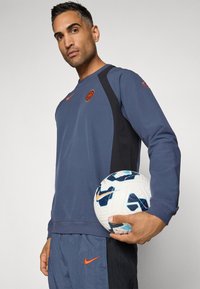 Felpa di colore blu scuro con pannelli laterali neri, dettagli arancioni e un logo. Il modello tiene in mano un pallone da calcio bianco e blu con un motivo.