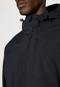 Veste à capuche noire avec fermeture éclair sur le devant, fabriquée en tissu léger. Dotée de cordons réglables sur la capuche et d'une texture lisse et mate.