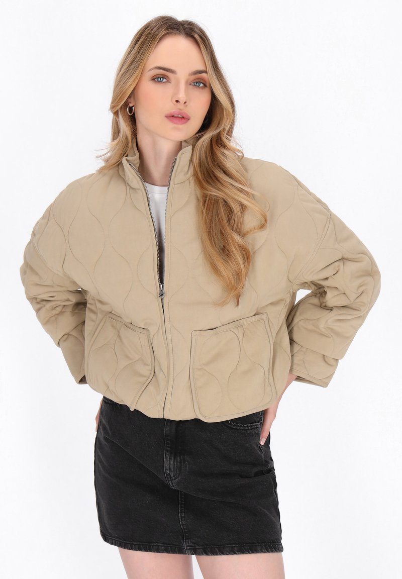 Beige gesteppte Jacke mit hohem Kragen, Reißverschluss und zwei Fronttaschen. Getragen über einem weißen Oberteil mit einem schwarzen Jeansrock.