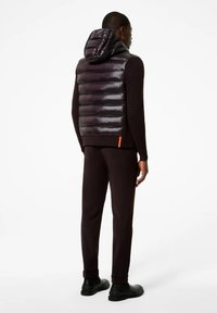 Gilet imbottito nero con cappuccio, dotato di una finitura lucida, motivo a righe orizzontali e un accento arancione vicino all'orlo, abbinato a pantaloni e scarpe scuri.