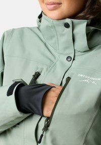 Jasnozielona kurtka typu soft-shell z wysokim kołnierzem, czarnymi akcentami, kieszeniami na zamek i nadrukowanym logo. Posiada regulowane mankiety.