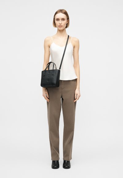 Femme portant un haut blanc sans manches, un pantalon marron, des bottines noires, portant un sac en cuir noir à bandoulière et un sac fourre-tout.