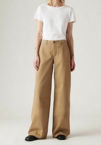Široké khaki kalhoty z bavlněné směsi, s knoflíkovým zapínáním a poutky na opasek; v kombinaci s jednoduchým bílým tričkem s krátkým rukávem.