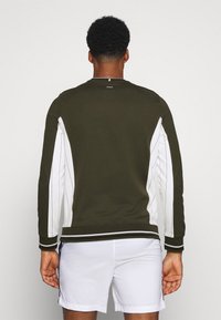 Sudadera color caqui con paneles laterales blancos y puños acanalados, con cuello redondo y un logo sutil en la parte posterior. Textura suave.