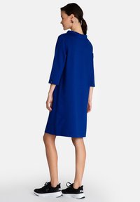 Blaues knielanges Kleid mit dreiviertelärmeln, flachem Kragen und seitlichen Akzenten. Kombiniert mit schwarzen Sportschuhen mit einer weißen Sohle.