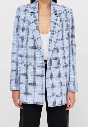 Donna che indossa una giacca a quadri azzurro chiaro con linee nere e bianche, bottoni dorati a forma di anello, top bianco e pantaloni neri.