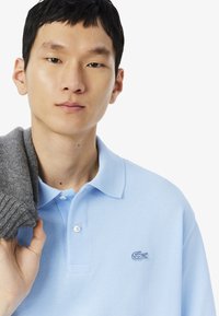 Ljusblå pikétröja med en texturerad väv, med en spetsig krage och en broderad logotyp på bröstet. Grå tröja hängande över axeln.