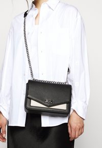 Borsa a tracolla nera in pelle con finitura texture, caratterizzata da un accento beige e una tracolla in catena argentata, mostrata su una camicia bianca.