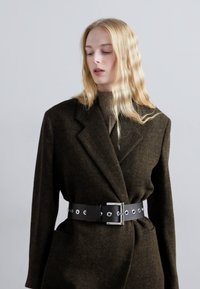 Ciemnozielony wełniany płaszcz z klapami, przewiązany czarnym skórzanym paskiem z metalowymi oczkami i prostokątną klamrą.