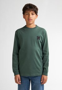 T-shirt verde a maniche lunghe in un morbido misto cotone, con collo a costine e una patch nera quadrata con un motivo circolare sul petto. Jeans in denim.