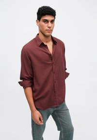 Tânăr stând cu brațele relaxate, îmbrăcat într-o cămașă maronie cu mânecile suflecate și blugi, pe un fundal alb simplu.