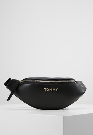 Sort læder bælletaske med guld lynlås og TOMMY logo i guld, placeret på en hvid piedestal mod en ensartet lys baggrund.