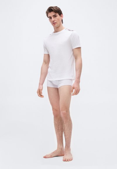 Νεαρός άνδρας όρθιος, ξυπόλυτος, φοράει λευκό εφαρμοστό μπλουζάκι και λευκά εσώρουχα boxer, μπροστά σε λευκό απλό φόντο.