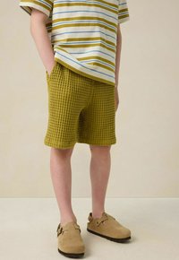 Chemise à manches courtes rayée en blanc, olive et bleu, associée à un short à carreaux texturé vert olive et des sabots beige.