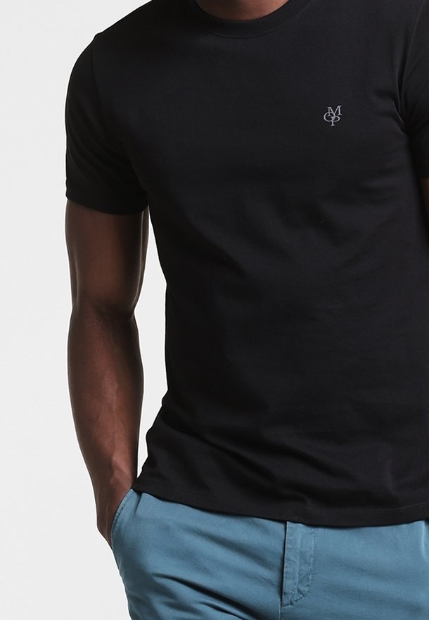 Schwarzes Baumwoll-T-Shirt mit Rundhalsausschnitt und kurzen Ärmeln. Verfügt über ein kleines graues Logo auf der linken Brust. Kombiniert mit hellblauen Hosen.