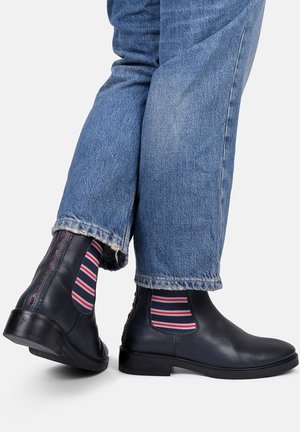 Czarne skórzane buty chelsea z elastycznymi bocznymi panelami, w połączeniu z niebieskimi dżinsami i paskowymi skarpetami w czerwono-białe paski. Gładkie wykończenie.