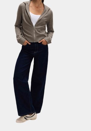 Person iført en lysebrun hættetrøje med lynlås, hvid tanktop, vidde ben mørk jeans og hvide sneakers med sorte striber.