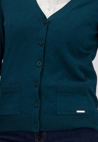 Teal gebreide cardigan met een V-hals, vijf knopen, twee opgestikte zakken, geribbelde zoom en een kleine metalen accent op de rechterzak.