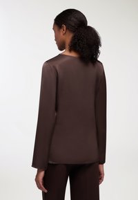 Maglia marrone a maniche lunghe con una texture liscia, scollatura rotonda e silhouette dritta, vista da dietro. Presenta un design minimale.