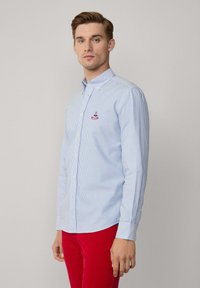 Camisa de botones a rayas azul claro y blanca con cuello, que presenta un pequeño logo bordado rojo en el pecho. Mangas largas y diseño ajustado.