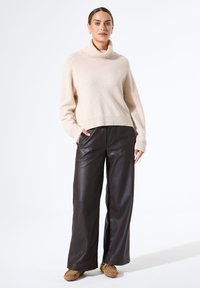 Beige Rollkragenpullover kombiniert mit schwarzen, weit geschnittenen Lederhosen, die einen lockeren Schnitt und eine glatte Textur aufweisen. Legere Schuhe runden den Look ab.