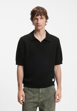 Jeune homme aux cheveux courts bruns portant un polo noir texturé à manches courtes et un pantalon vert à motifs sur un fond uni.