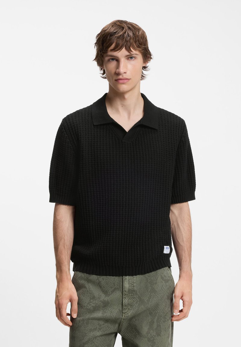 Jeune homme aux cheveux courts bruns portant un polo noir texturé à manches courtes et un pantalon vert à motifs sur un fond uni.