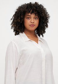 Camicia bianca button-up in tessuto leggero, con colletto classico, maniche lunghe e una vestibilità rilassata con una texture sottile.