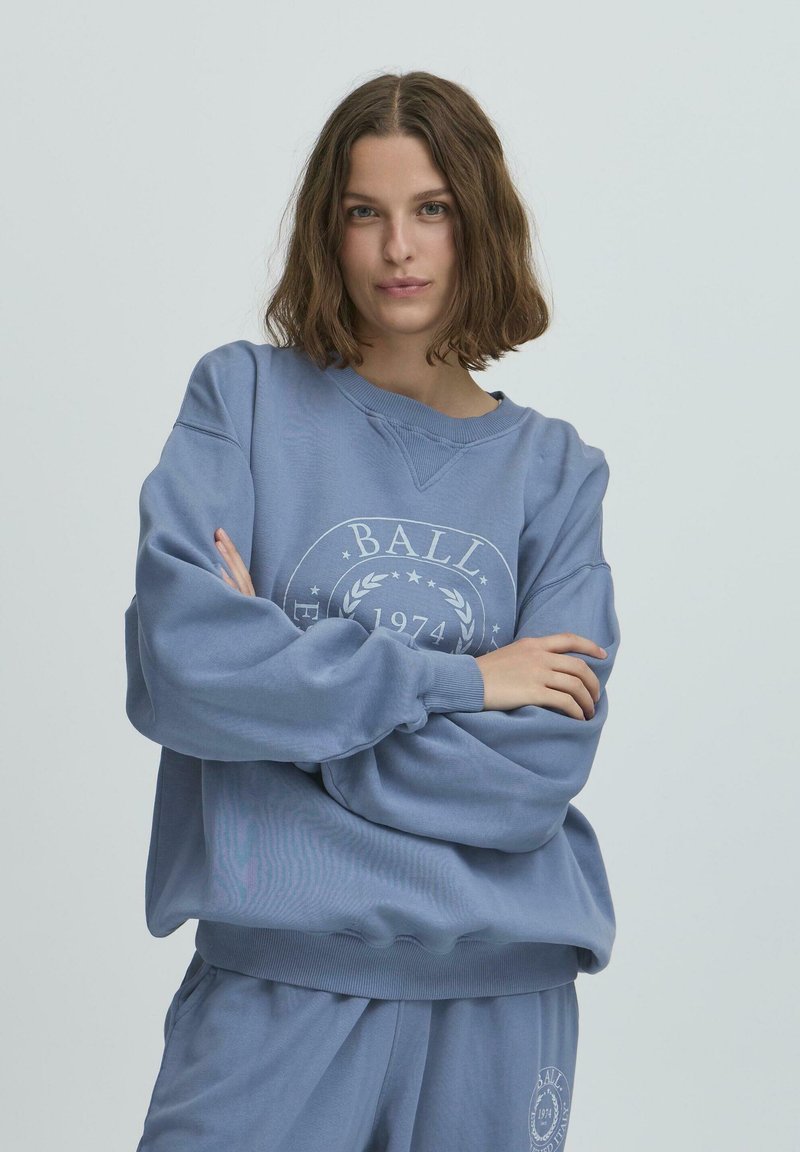 Gaiši zils džemperis, kas izgatavots no mīksta materiāla, ar apaļu grafisko logotipu un tekstu, ribotiem piedurknēm un brīvu piegriezumu.