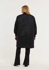 Vrouw met lang blond haar, gekleed in een lange zwarte gewatteerde jas, zwarte flarebroek en zwarte puntige hakken, staande tegen een effen achtergrond.