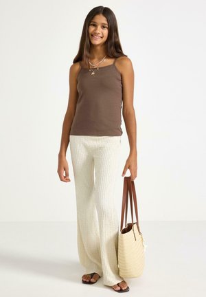 Hymyilevä tyttö, jolla on pitkä ruskea tukka, pukeutuneena ruskeaan tankkitoppiin, teksturoituihin valkoisiin housuihin, mustiin sandaaleihin ja hän pitää kädessään vaaleanruskeaa kudottua olkalaukkua, jossa on ruskeat hihnat.