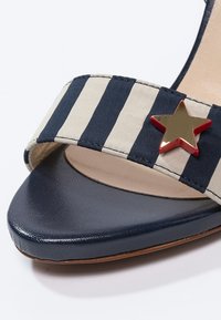 Sandale à talons hauts bleu marine avec une bride en tissu rayé crème et bleu marine, accentuée d'une embellissement en étoile dorée et rouge.